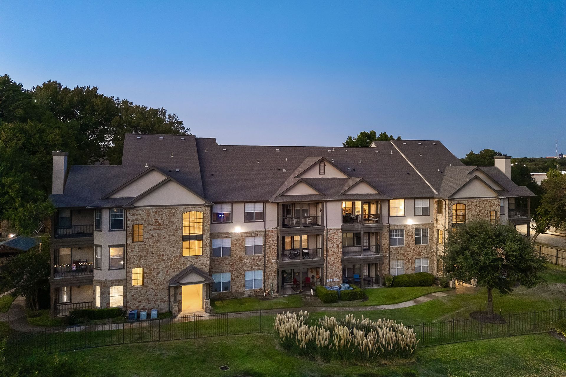 Large stone apartment building at dusk with lit windows, trees, and a landscaped lawn at Marquis at Bellaire Ranch in Fort Worth, TX