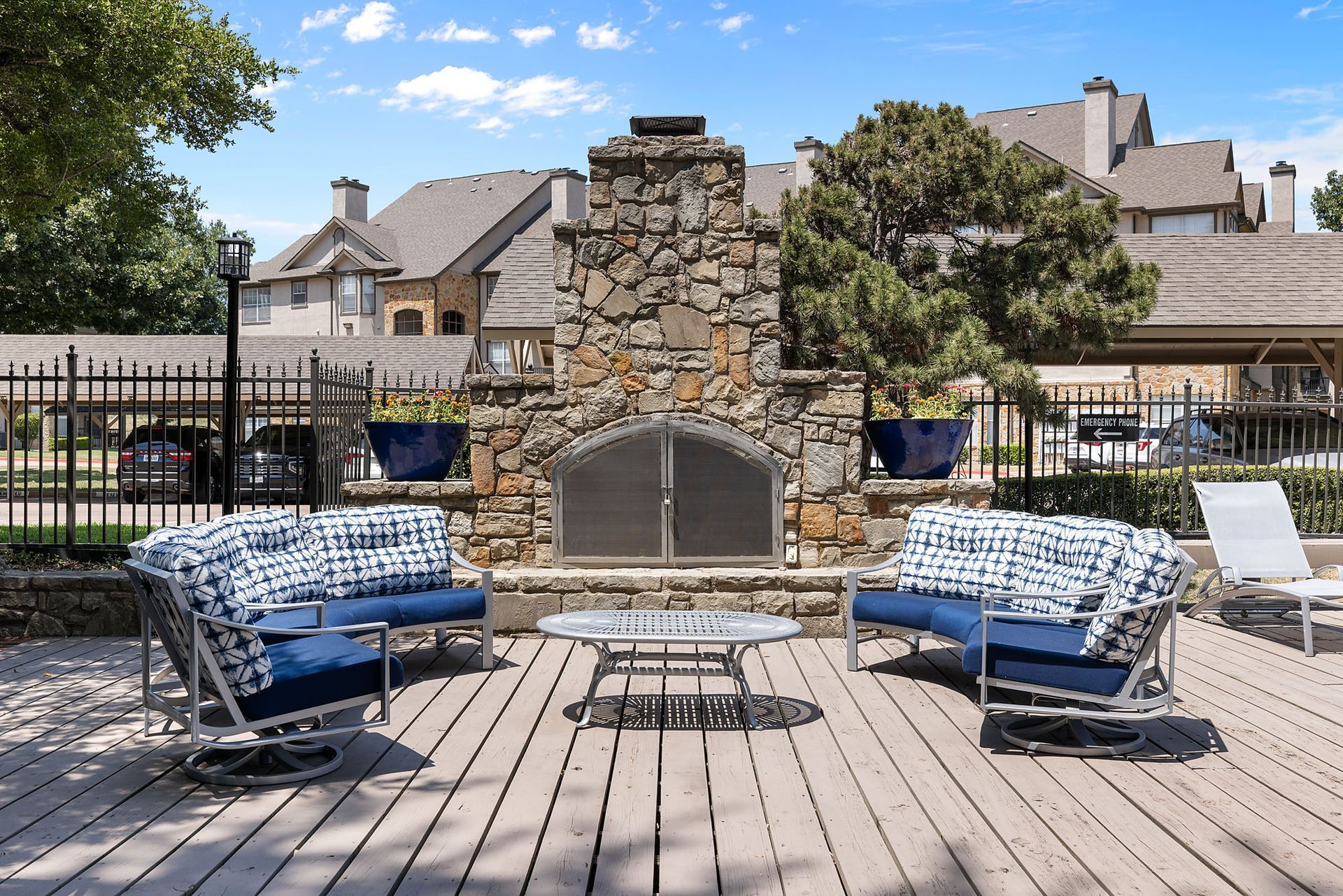 Patio with stone fireplace, blue chairs, and table on a wooden deck under a bright sky at Marquis at Bellaire Ranch in Fort Worth, TX.