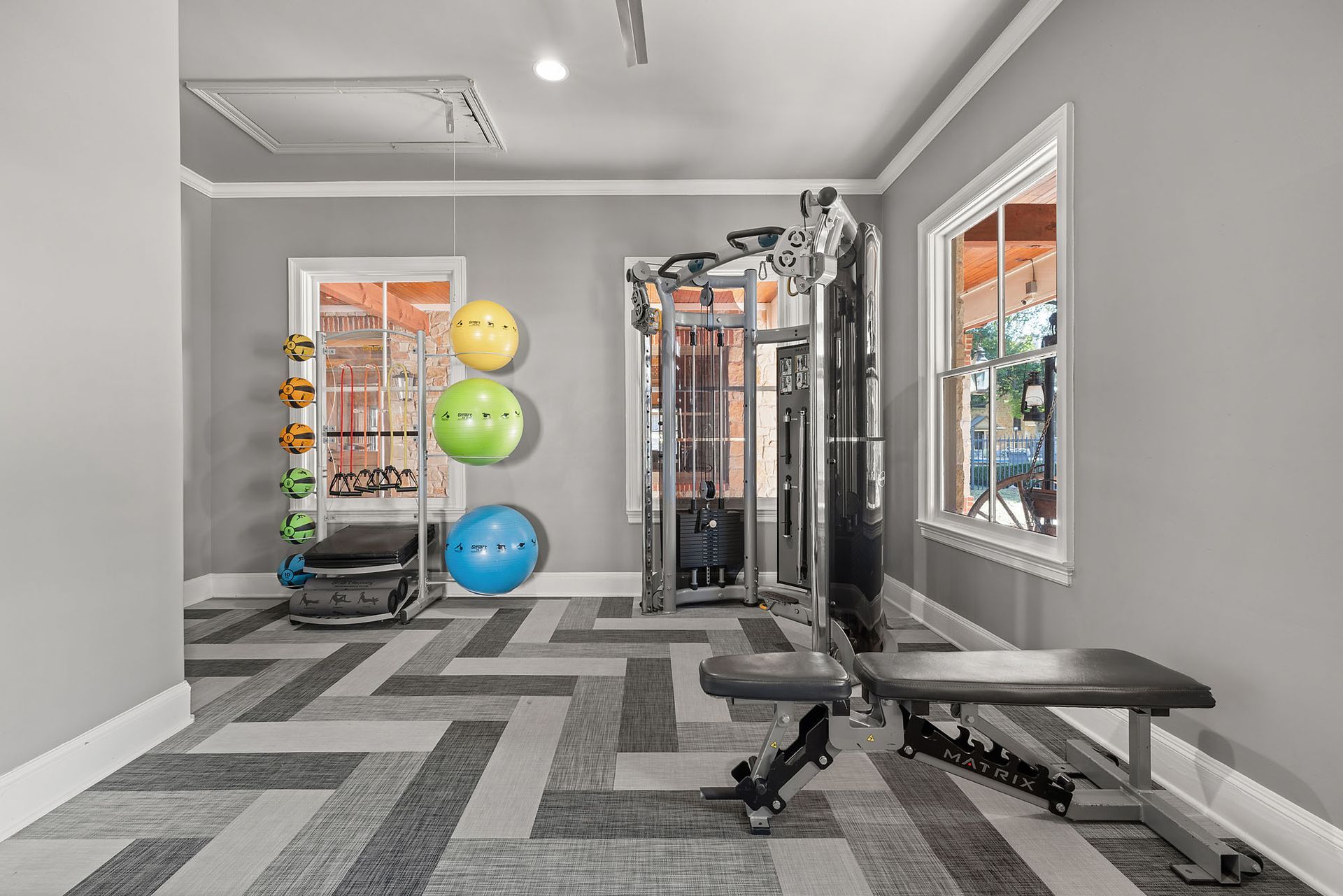 Small gray gym with cable machine, bench, stability balls, and checkerboard carpet floor at Marquis at Bellaire Ranch in Fort Worth, TX