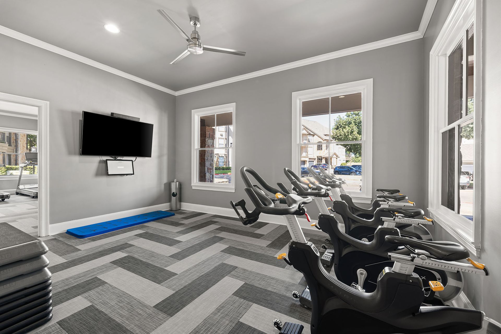 Modern gym with treadmills, wall-mounted TV, gray chevron carpet, and large windows at Marquis at Bellaire Ranch in Fort Worth, TX