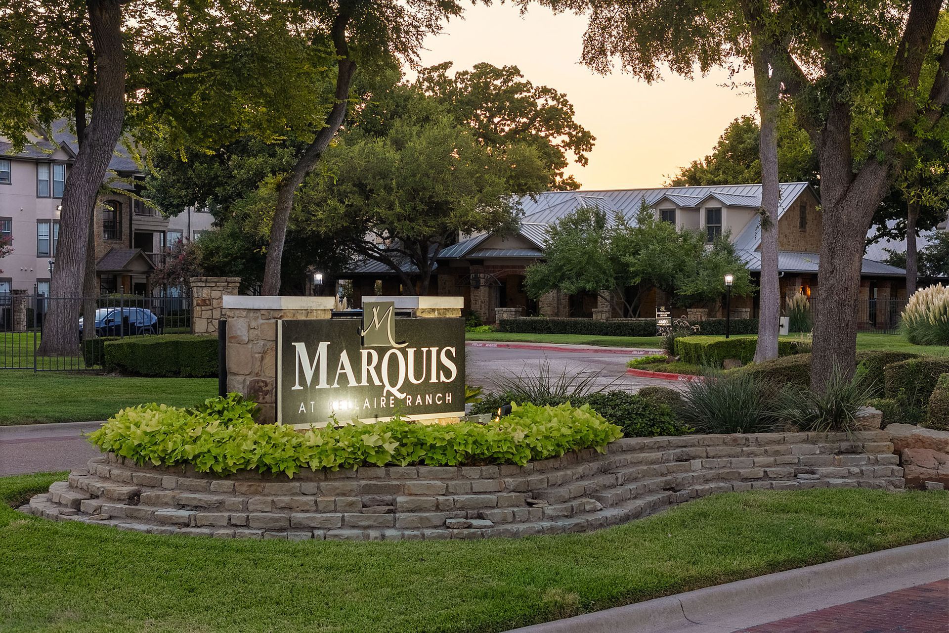Marquis apartment community sign in a landscaped courtyard at dusk, with trees and buildings behind it at Marquis at Bellaire Ranch in Fort Worth, TX