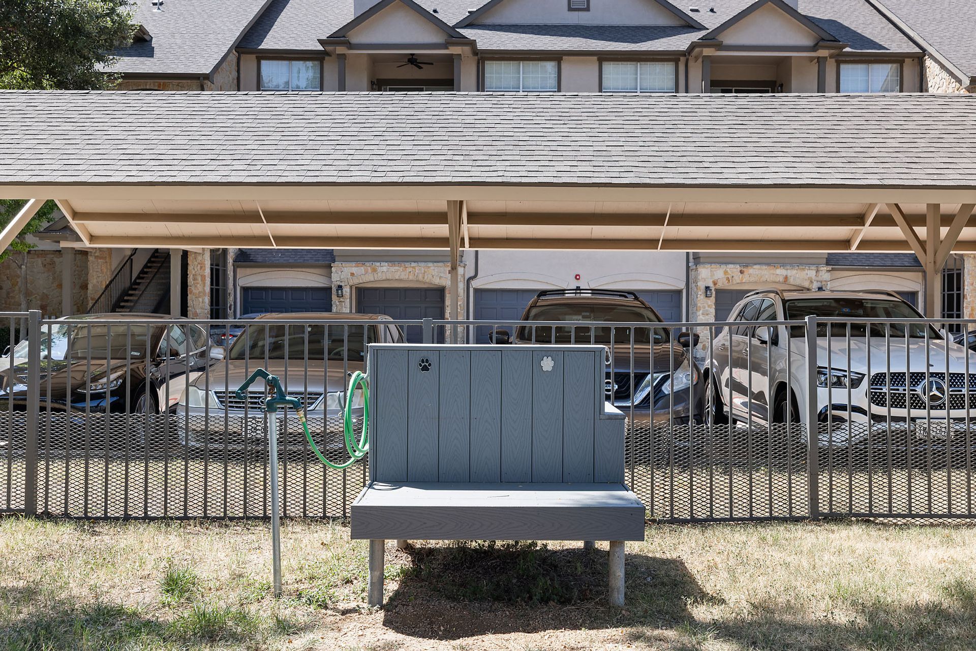 Gray bench in front of a fenced apartment parking area with cars under a covered structure at Marquis at Bellaire Ranch in Fort Worth, TX.