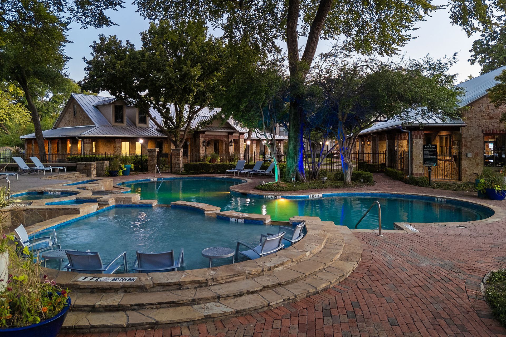 Outdoor resort pool with blue water, stone patio, and lit cottages at dusk at Marquis at Bellaire Ranch in Fort Worth, TX