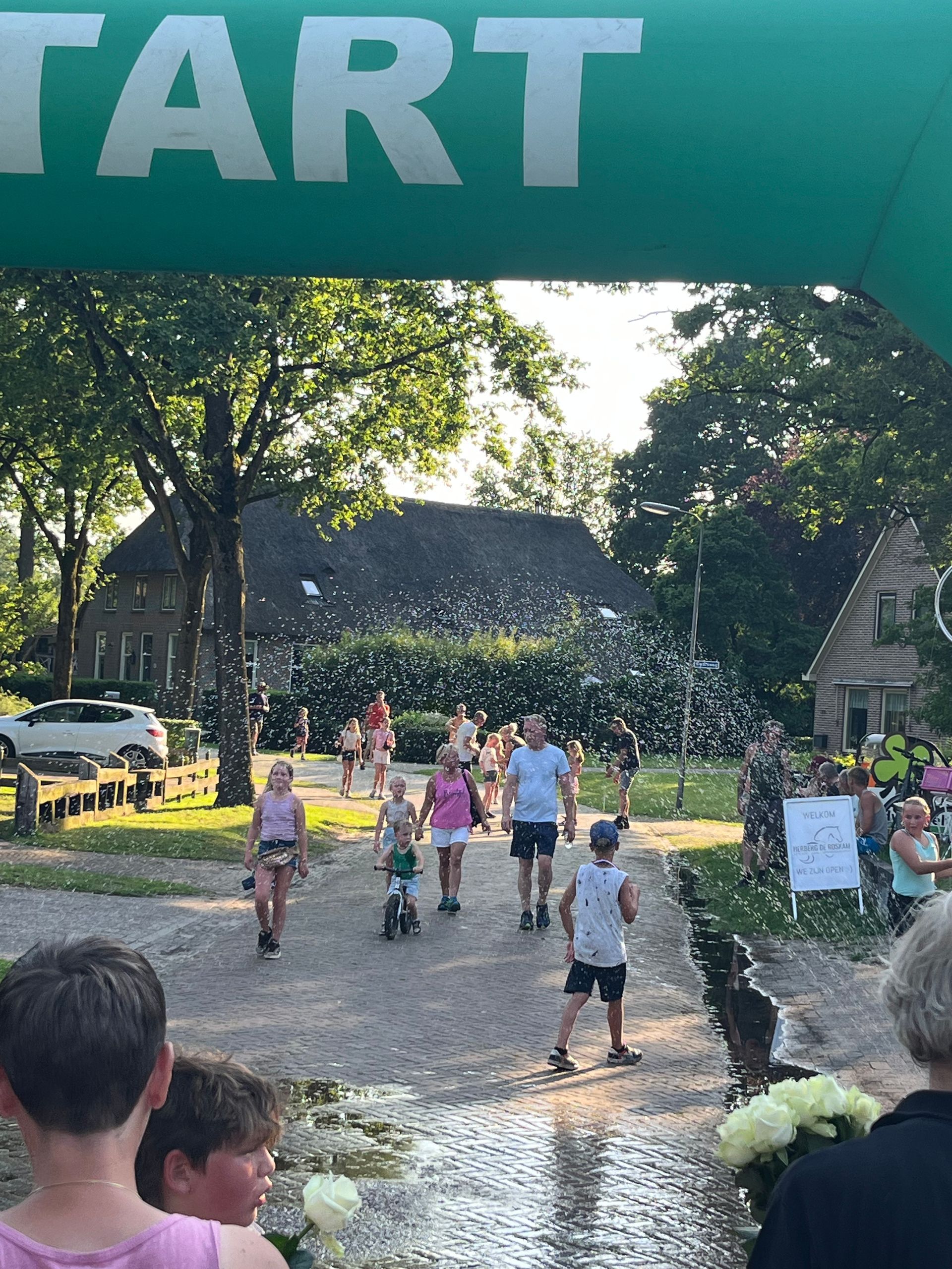 Hardlopers en kinderen onder een groene START-boog bij 4daagse, met opspattend water langs het pad.