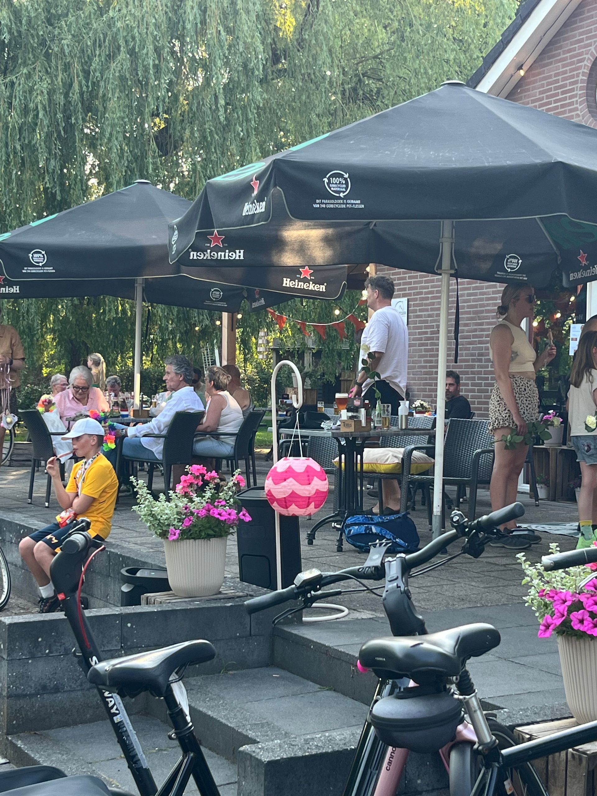Terras van een café met zwarte parasols, mensen aan tafels en roze bloemen bij de stenen trappen.