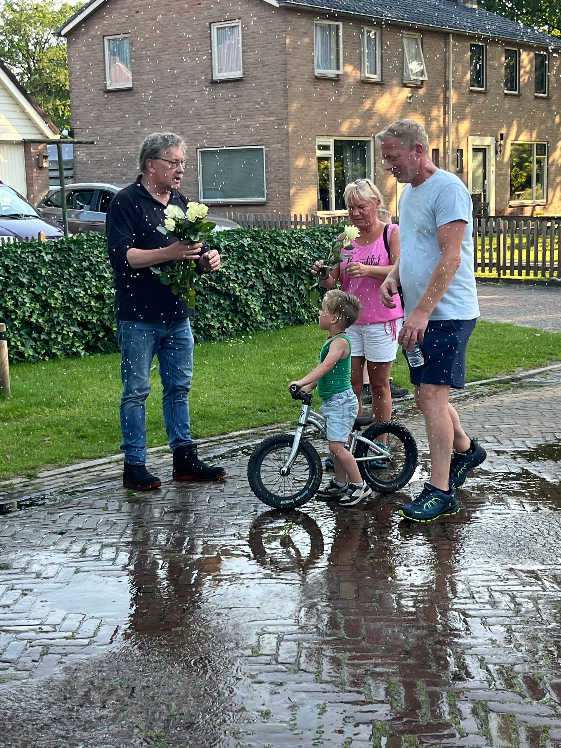 Mensen staan ​​op een natte oprit, een van hen houdt een kinderfietsje vast, voor een bakstenen huis.