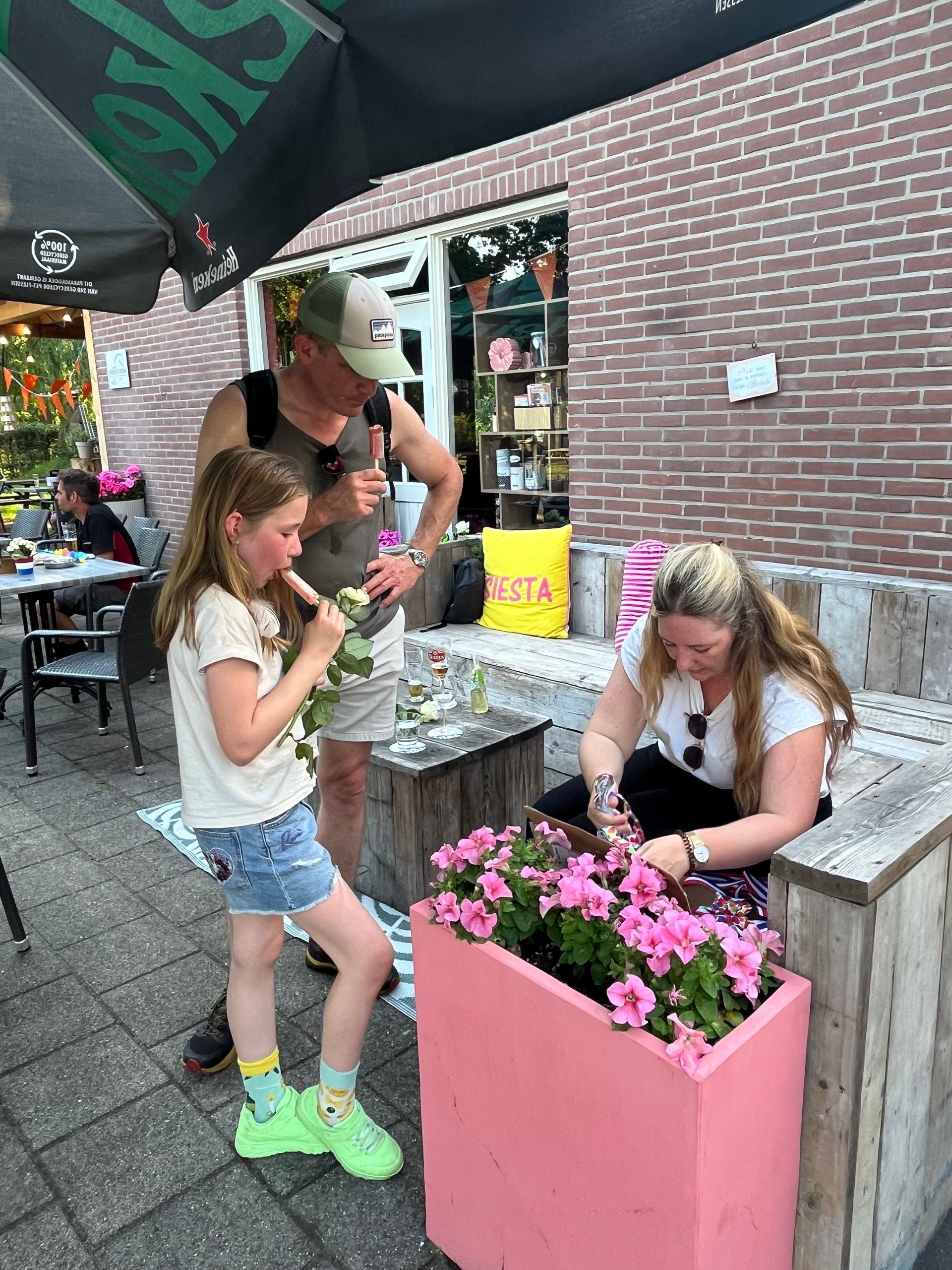 Twee meisjes verzorgen roze bloemen in een bloembak naast een bakstenen gebouw, met een vrouw in de buurt op het terras