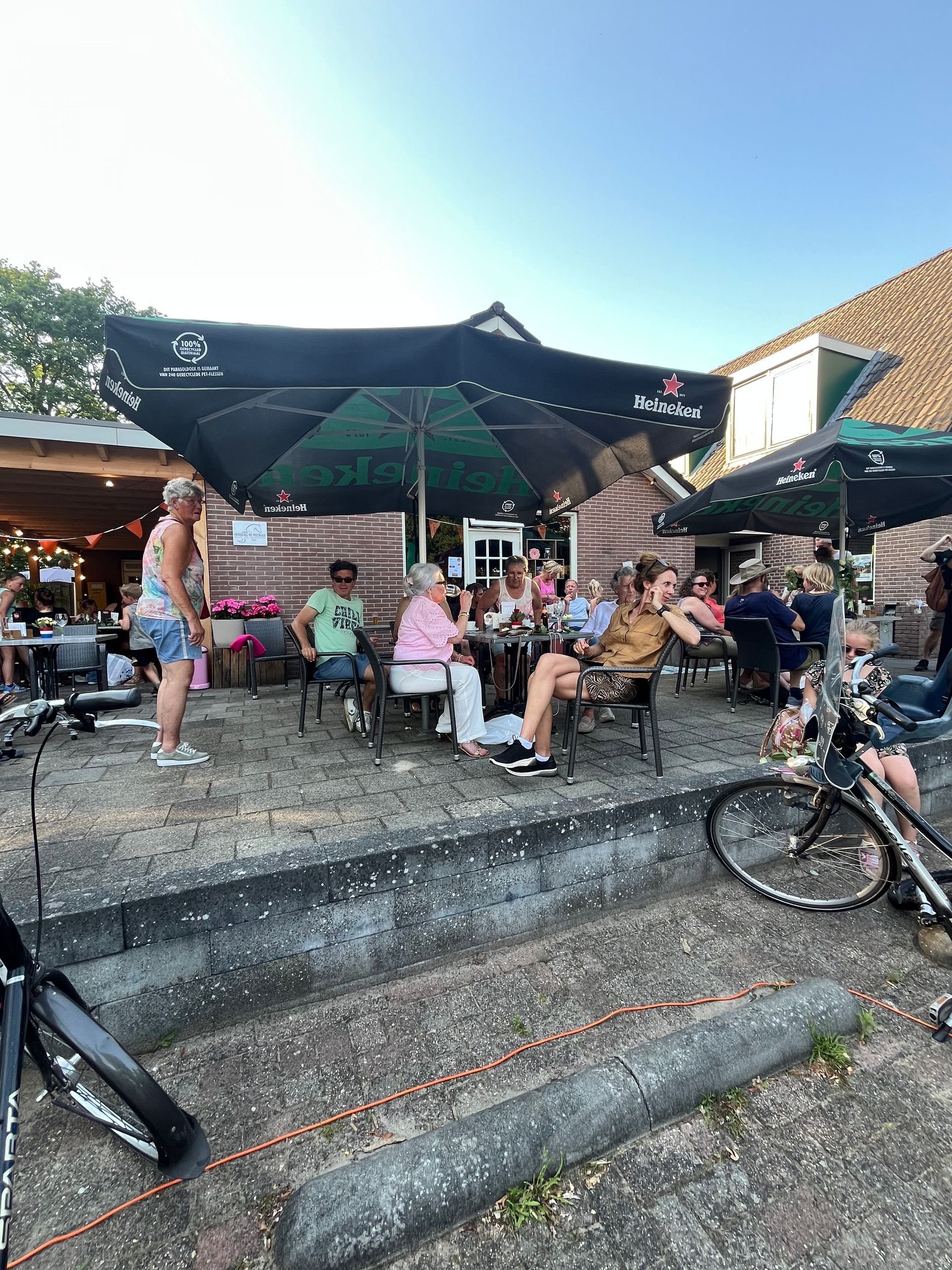Terras van een café in de buitenlucht met mensen die onder zwarte parasols zitten, met fietsen op de voorgrond.