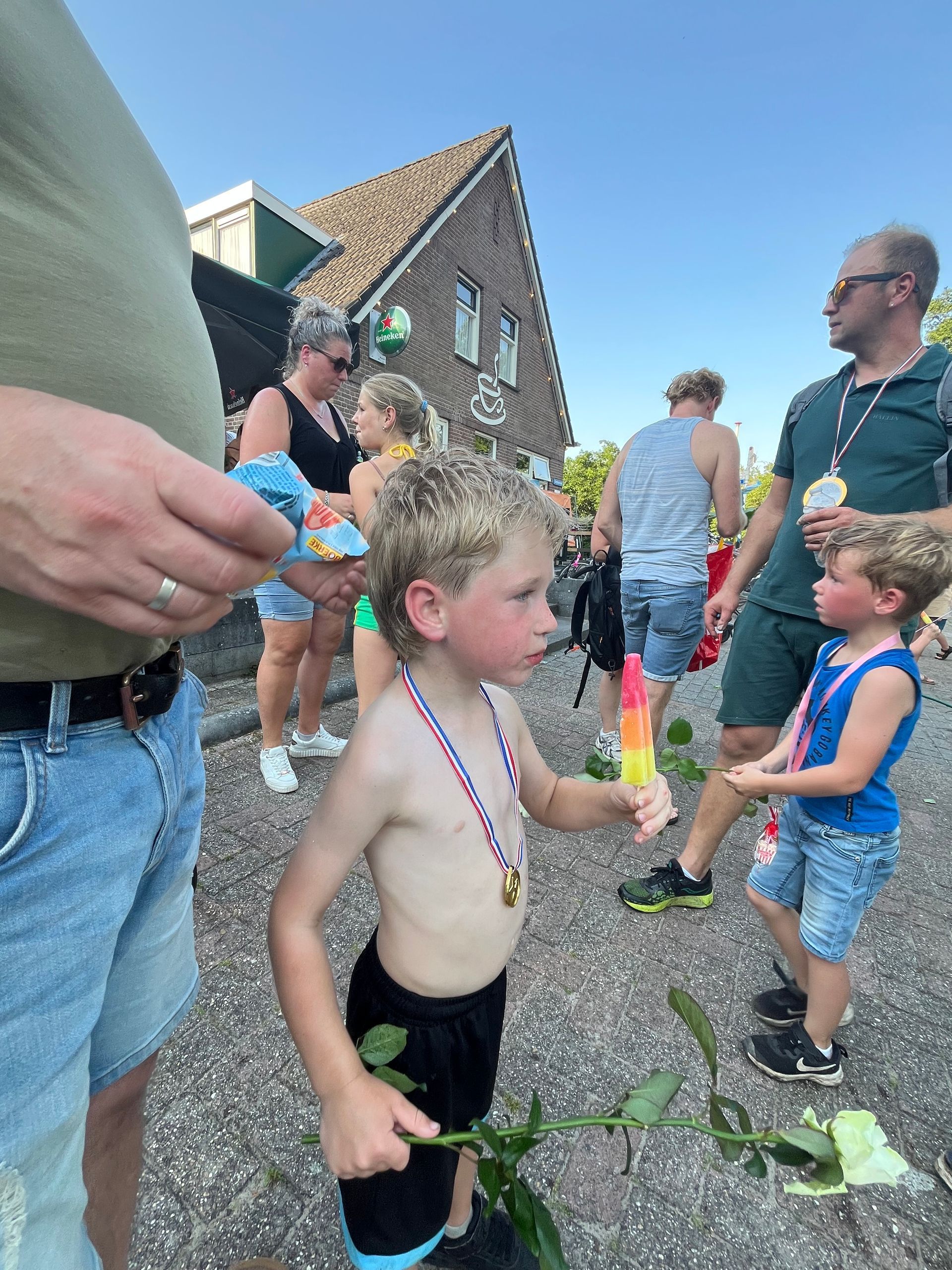 Kinderen spelen buiten in de buurt van een huis, waarbij een van de kinderen een kralenketting draagt ​​en een bloemsteel vasthoudt.