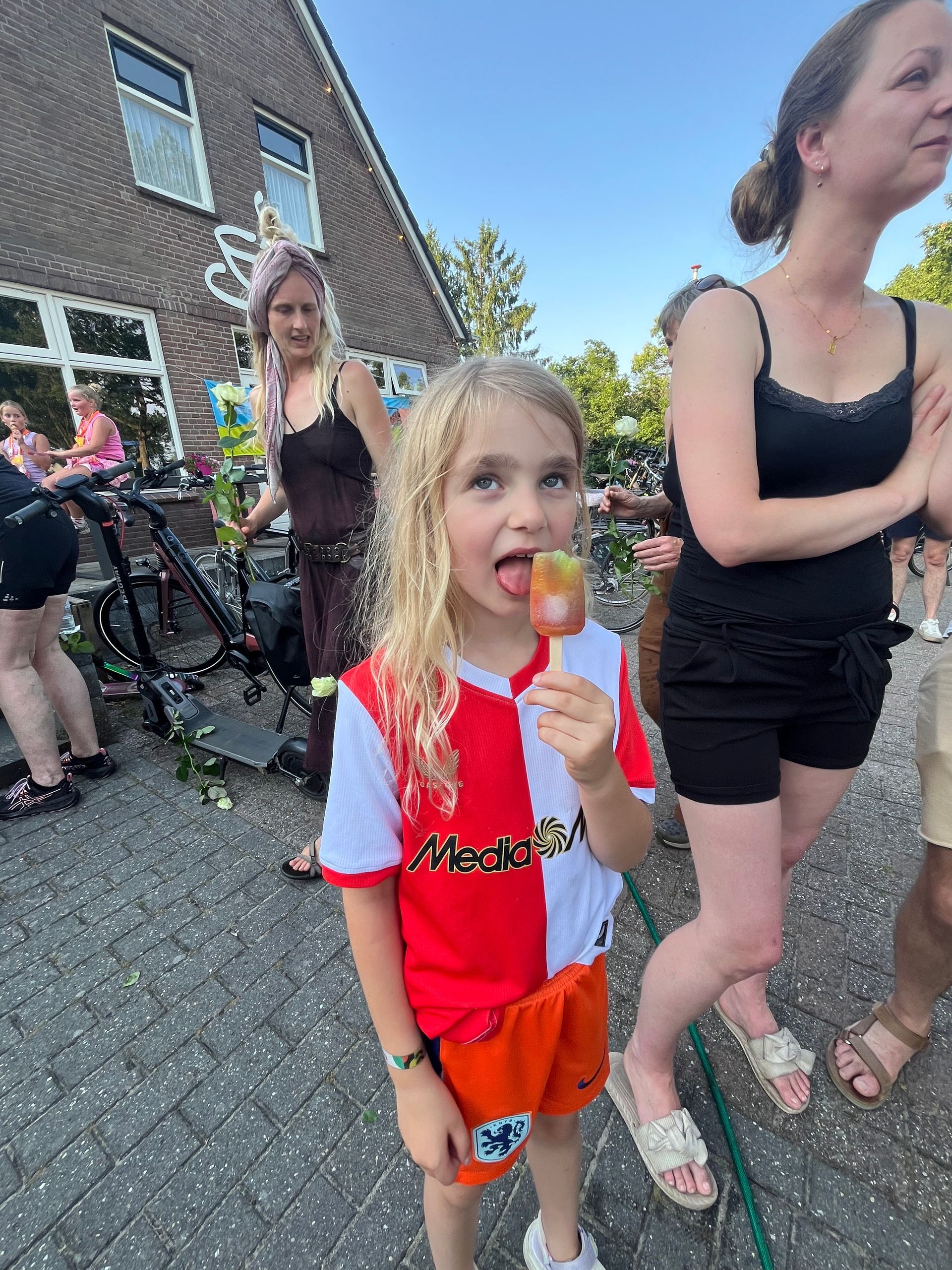 Kind in rood-wit shirt eet een sinaasappelijsje tijdens een buitenbijeenkomst.