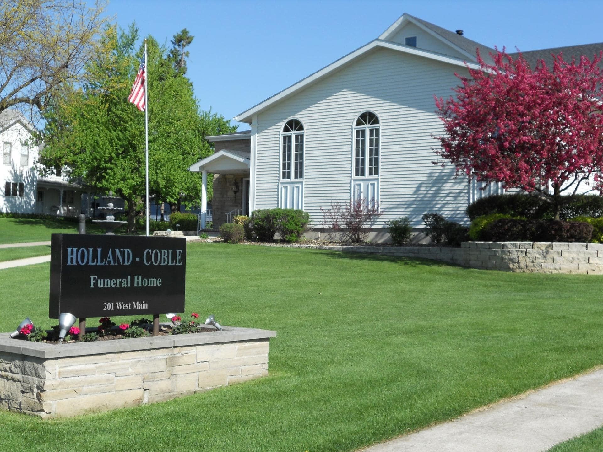 Holland-College Funeral Home sign in front of a white building with an American flag and blooming trees