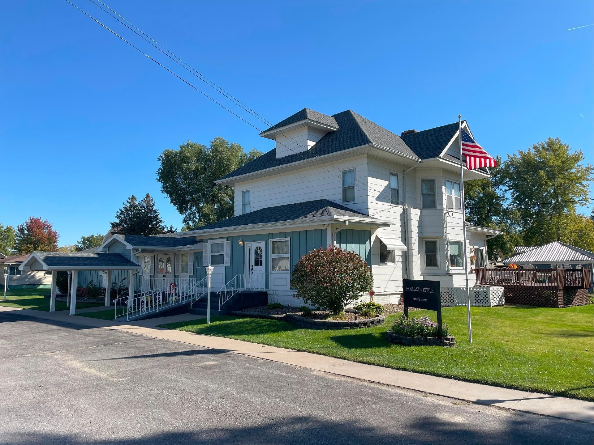 White Victorian-style house with a porch, American flags, and a grassy yard under a clear blue sky