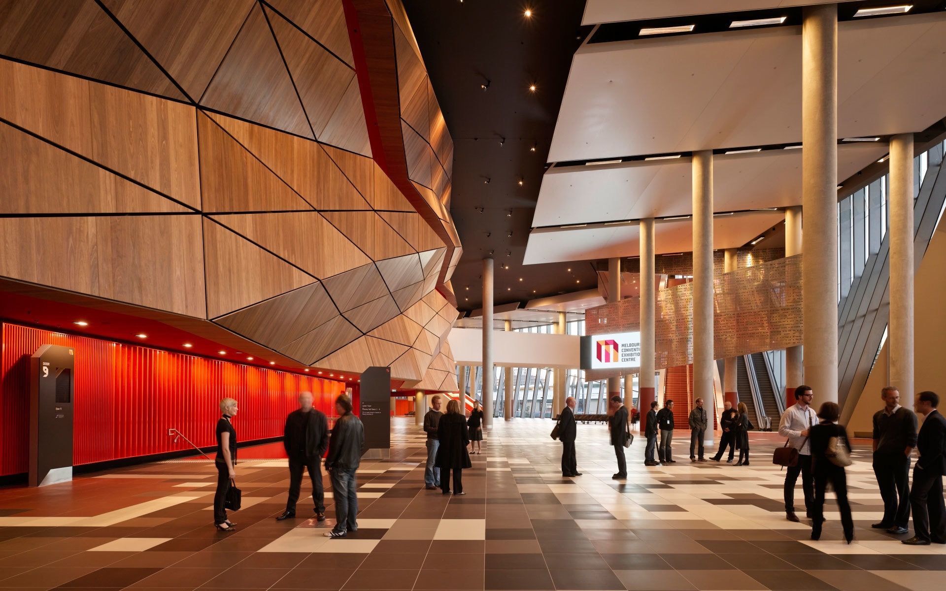 Modern indoor corridor with red walls, tall columns, and people walking in a brightly lit venue