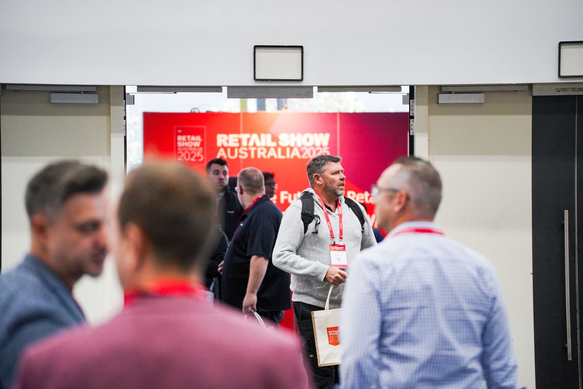 Trade show hallway with attendees wearing red lanyards and a red “Retail Show Australia” banner.