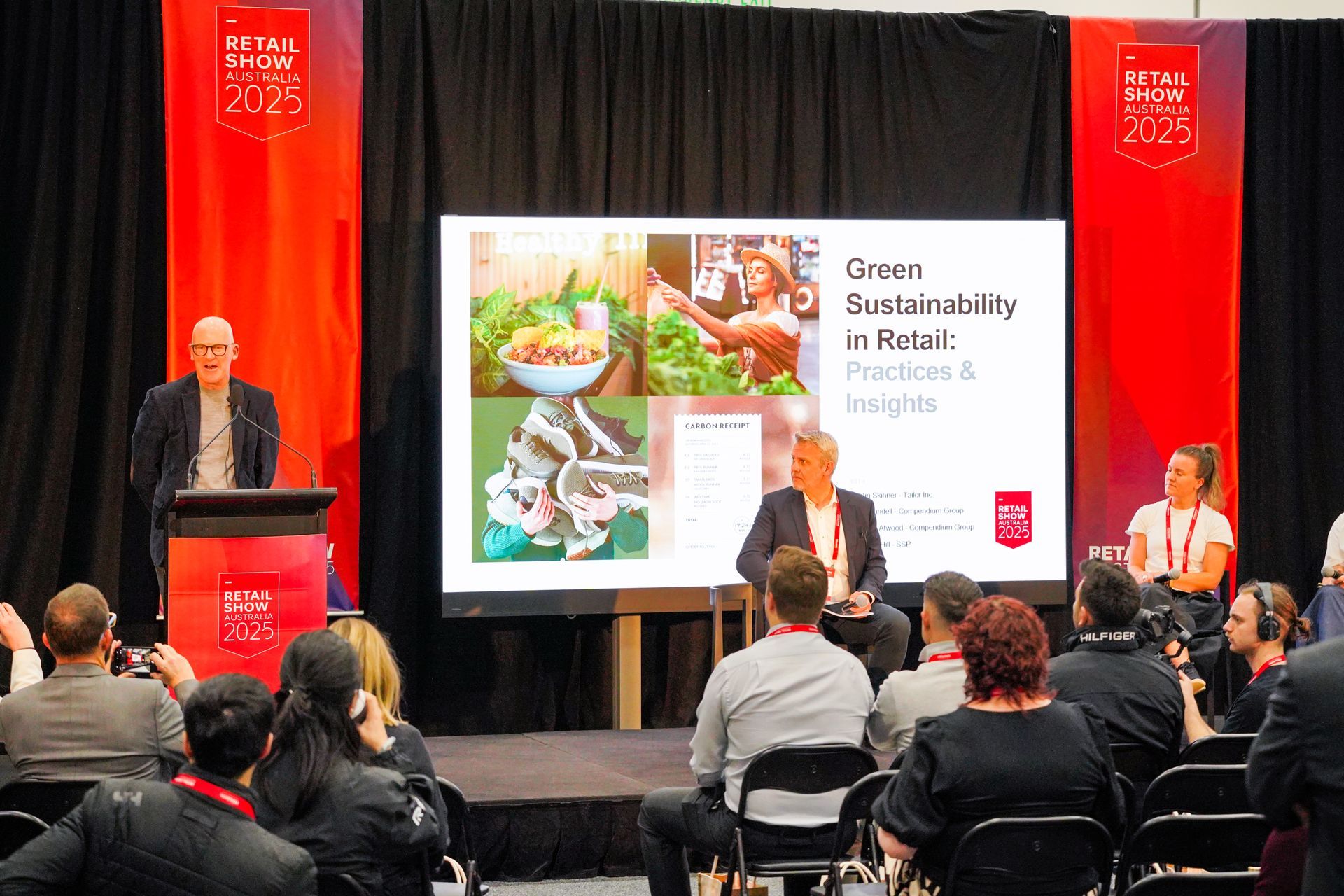 Panel discussion on stage with audience, red curtains, and presentation screen in the background