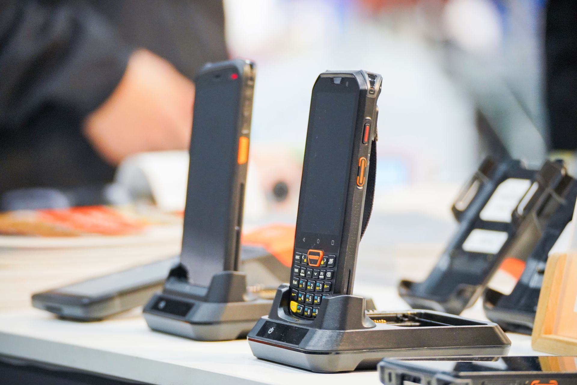Rugged handheld radios on charging docks on a tabletop, with blurred people in the background