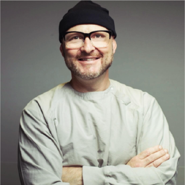 Smiling man in glasses and black cap, arms crossed, wearing a light gray shirt against a gray background