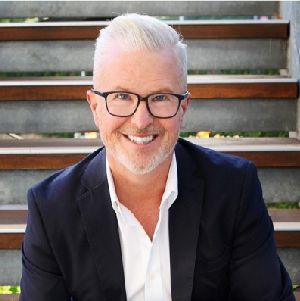 Smiling person with white hair and glasses in a dark blazer, seated on outdoor steps.