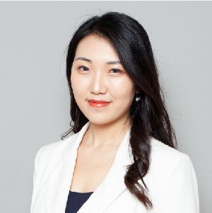 Portrait of a woman in a white blazer against a gray background, smiling softly.
