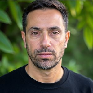 Head-and-shoulders portrait of a man in a black shirt against a blurred green background