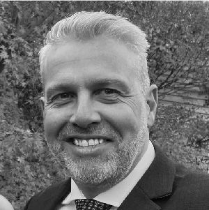 Smiling man in a suit outdoors, with trees in the background.