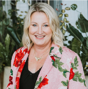 Smiling blonde woman in a pink floral blazer against a leafy indoor backdrop