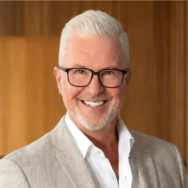 Smiling man with white hair and glasses in a light gray blazer against a wood-paneled background
