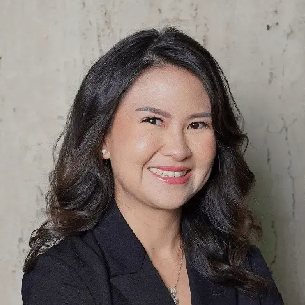 Smiling woman with wavy dark hair in a black blazer against a light stone background