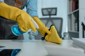Worker in yellow gloves disinfects a desk with a blue spray bottle in an office.