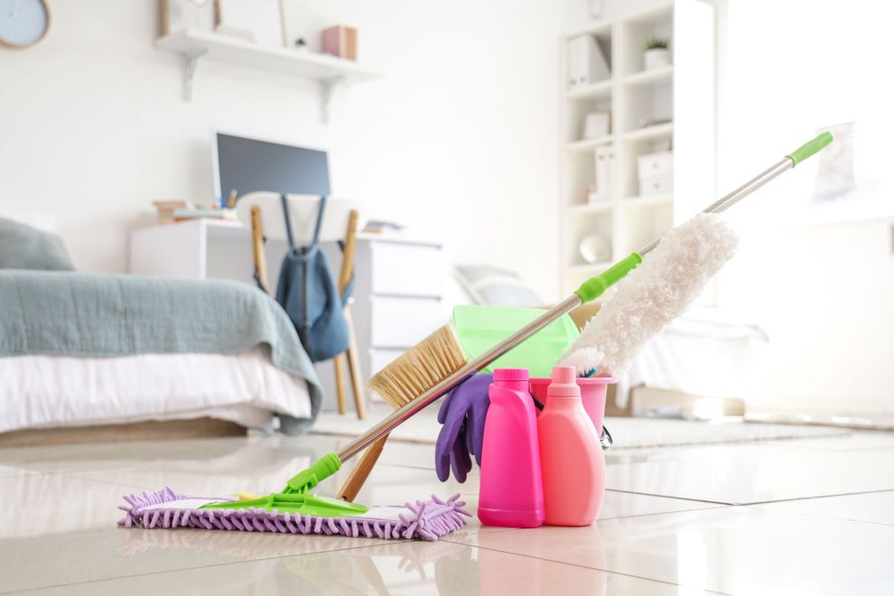 Cleaning supplies on a floor in a bright bedroom, with a mop and spray bottles near a bed.