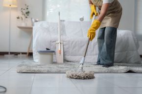 Person mopping a bright living room floor beside a white sofa and lamp