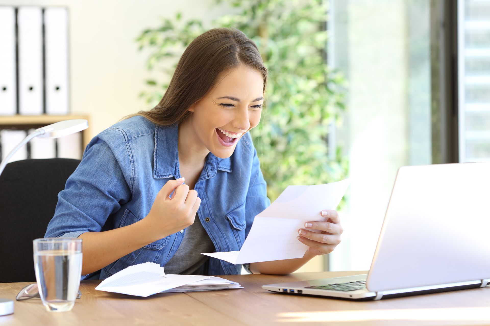 A woman is sitting at a desk holding a piece of paper in front of a laptop computer.