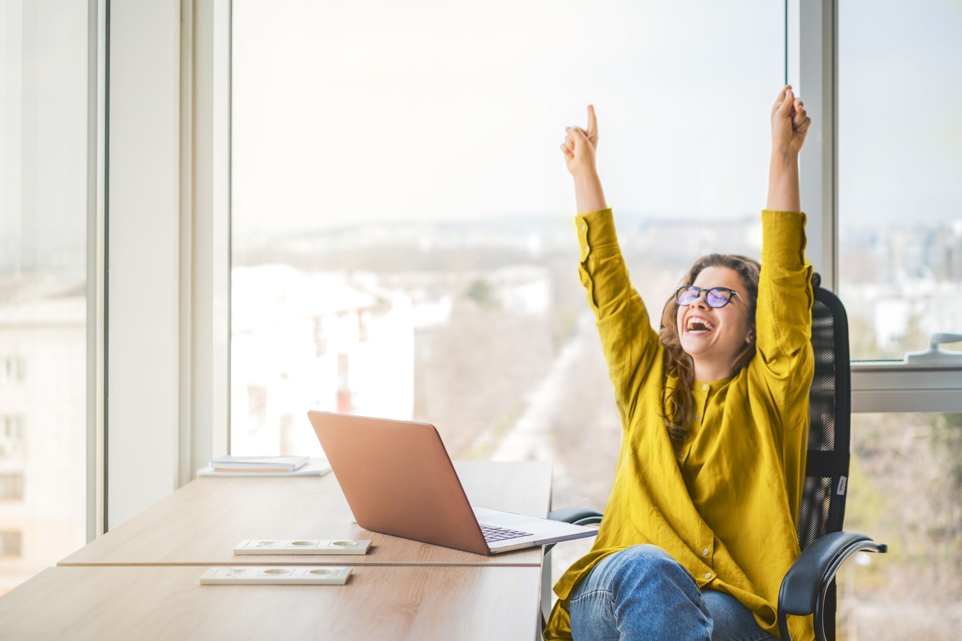 A woman is sitting at a desk with her arms in the air in front of a laptop computer.