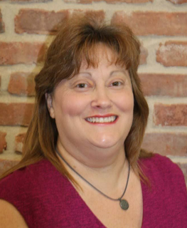 Smiling woman in a magenta top and necklace standing against a brick wall.