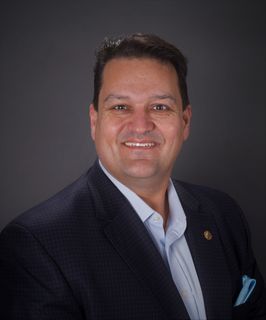 Professional headshot of a smiling man in a dark suit and white shirt against a gray background