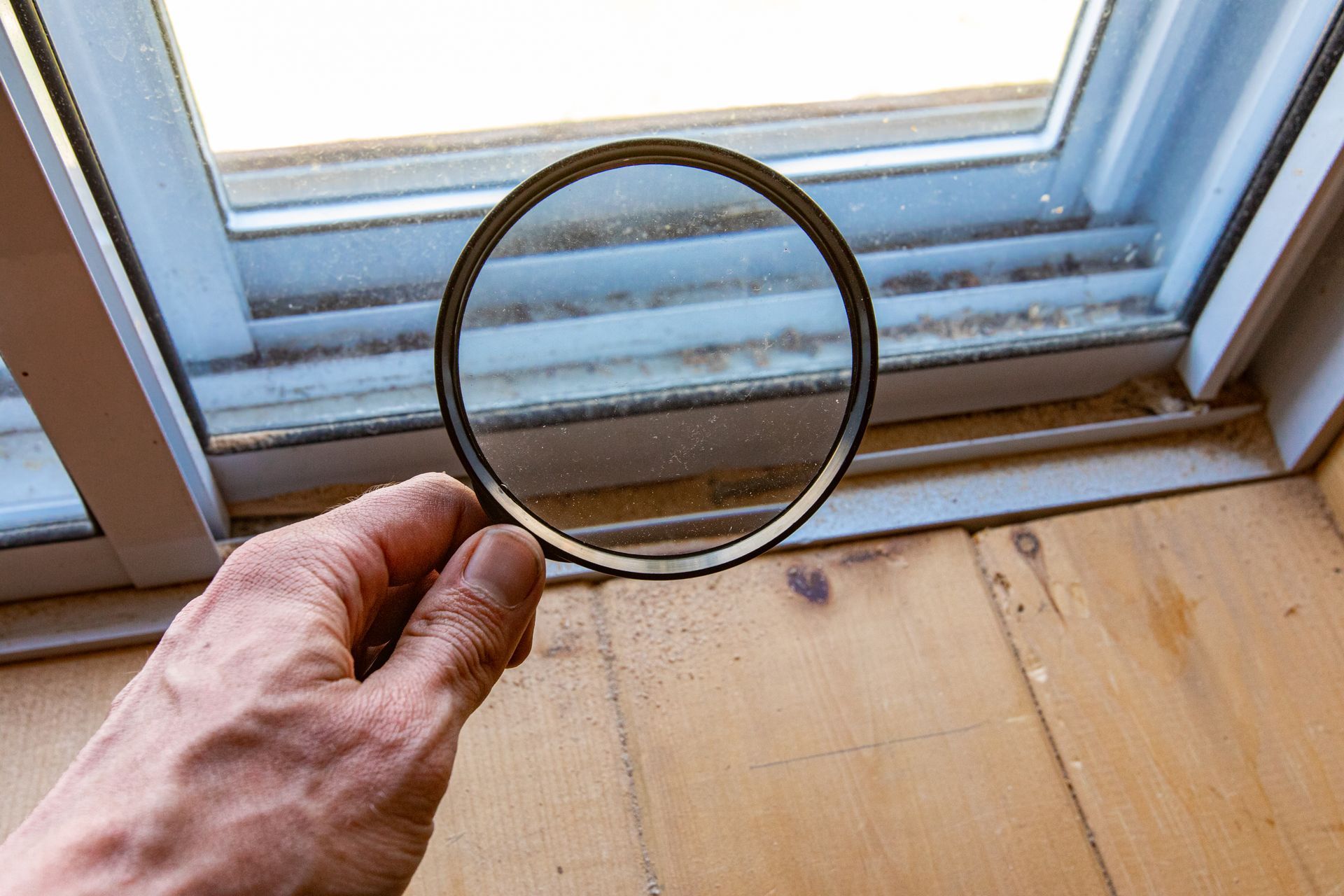 Hand holding a magnifying glass near a dusty window sill and wooden floorboards