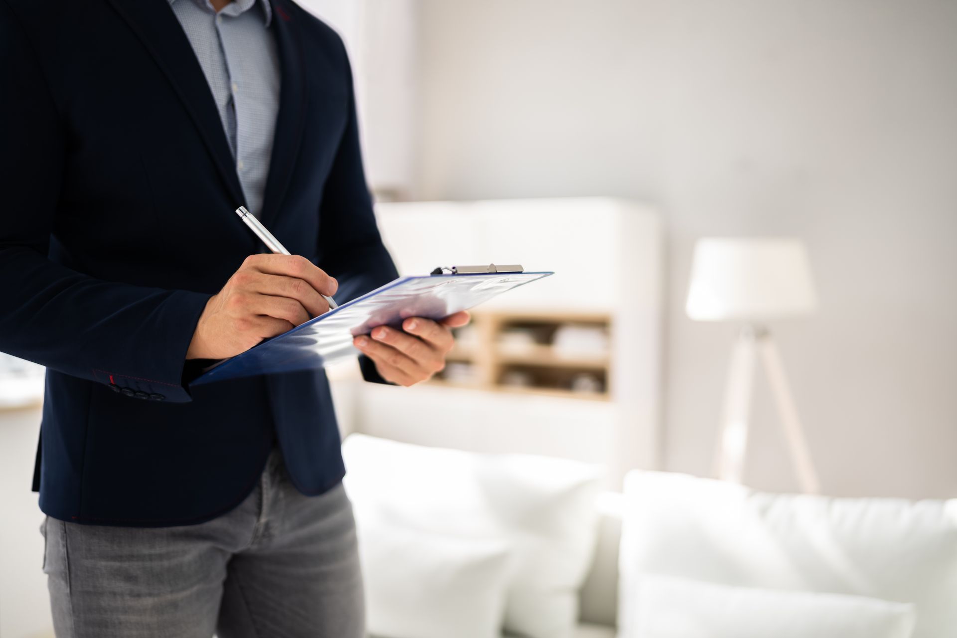 Person in a navy blazer writing on a clipboard in a bright bedroom with white furniture