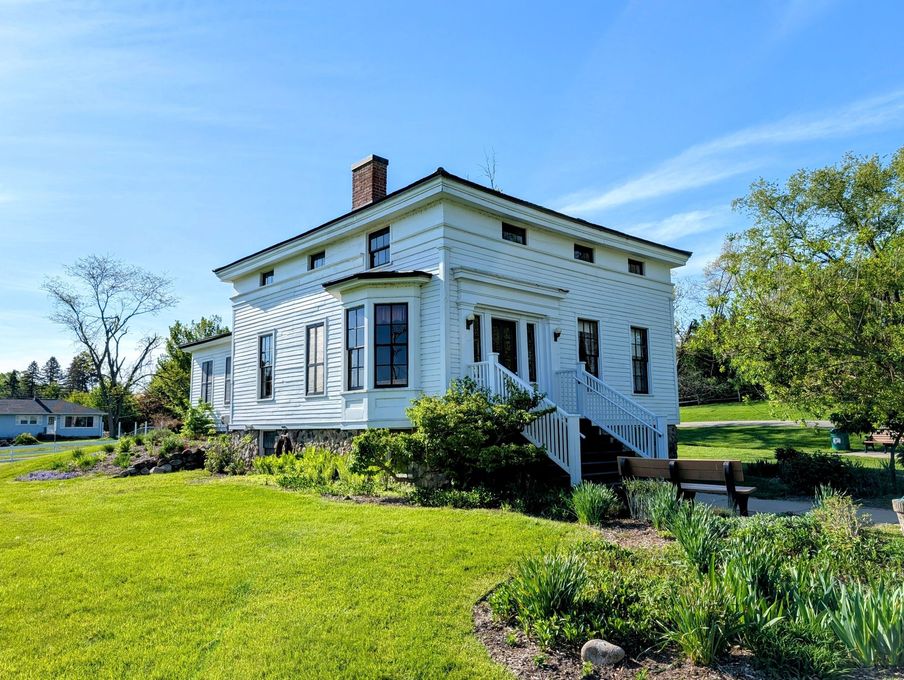 White farmhouse with blue trim on a grassy hill under a clear blue sky, surrounded by trees and garden plants
