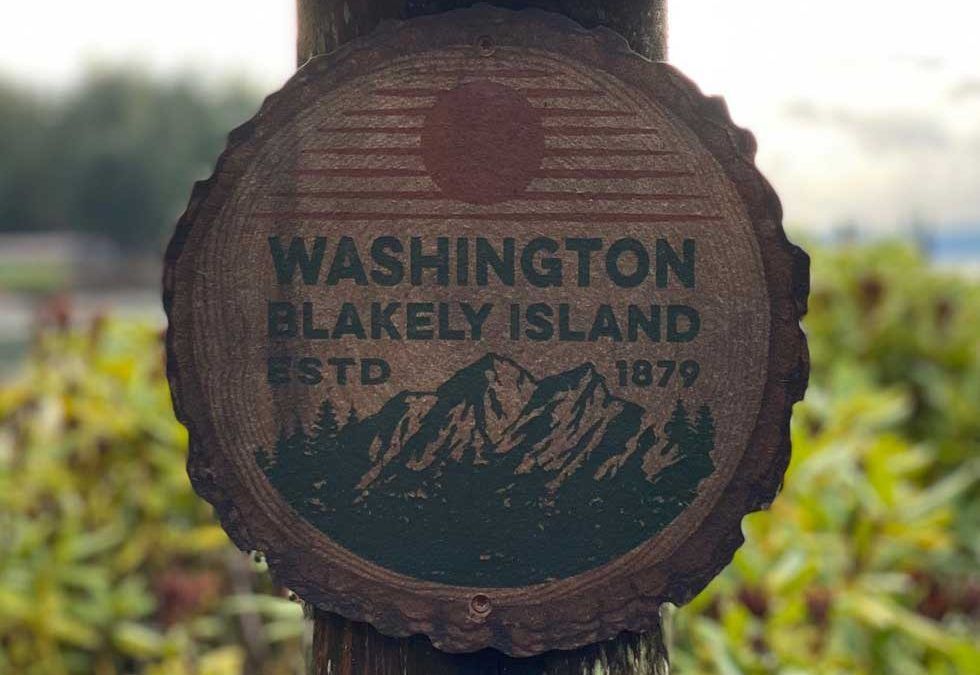 Wooden sign for Blakely Island, Washington, with text and mountain/forest graphic, blurred background.