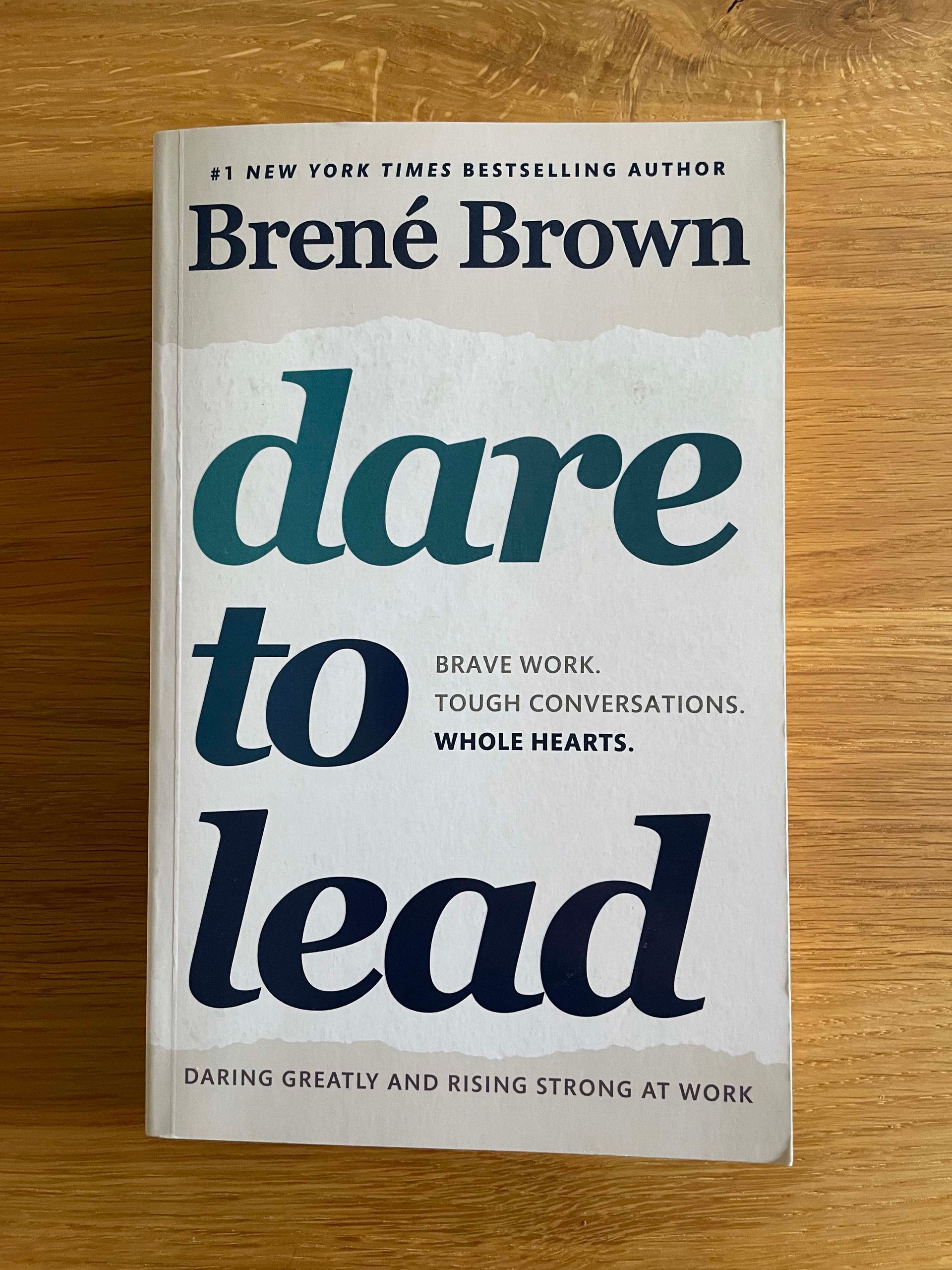 Foto des englischen Buchcovers von Dare to Lead von Brené Brown auf einer Holzoberfläche. Das Buchcover ist weiß and grau mit dunkelblauer Schrift mit Farbverlauf. Der Untertitel lautet: brave work, tough conversations, whole hearts.