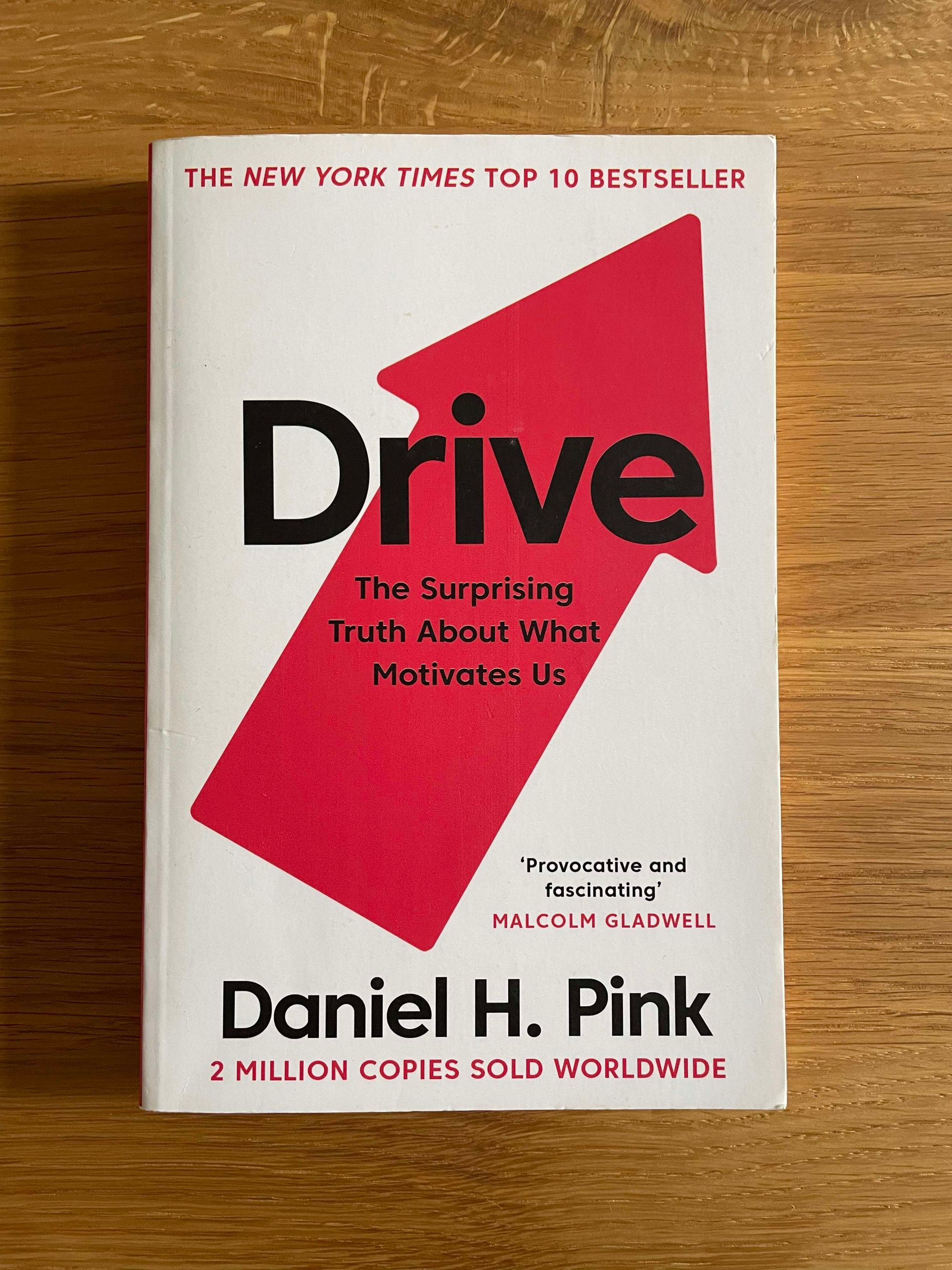 Foto des englischen Buchcovers von Drive von Daniel H. Pink auf einer Holzoberfläche. Das Buchcover ist weiß mit einem roten Pfeil nach oben. Die Schrift ist schwarz und rot. Der Untertitel lautet: The surprising truth about what motivates us.