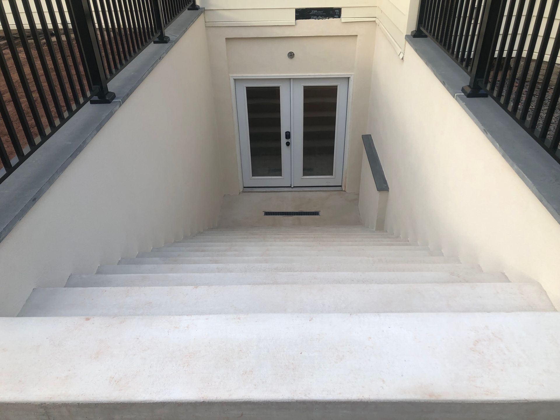 White concrete stairs leading down to a set of double glass doors framed by light-colored walls and black metal railings.