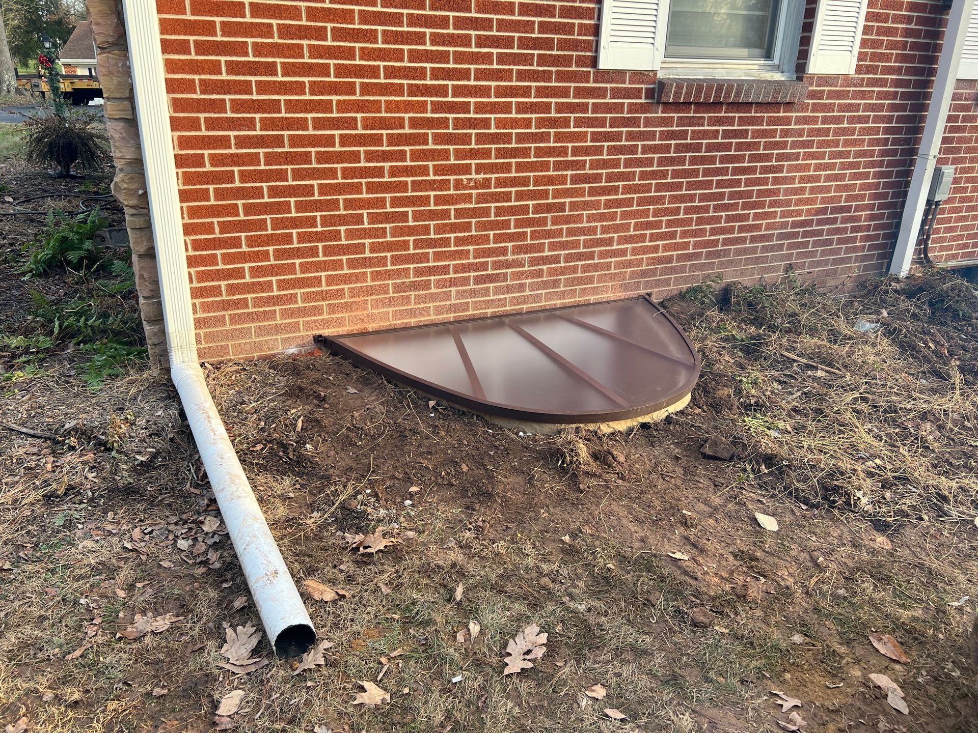 Basement window well beside red brick house with brown metal cover and a downspout pipe nearby