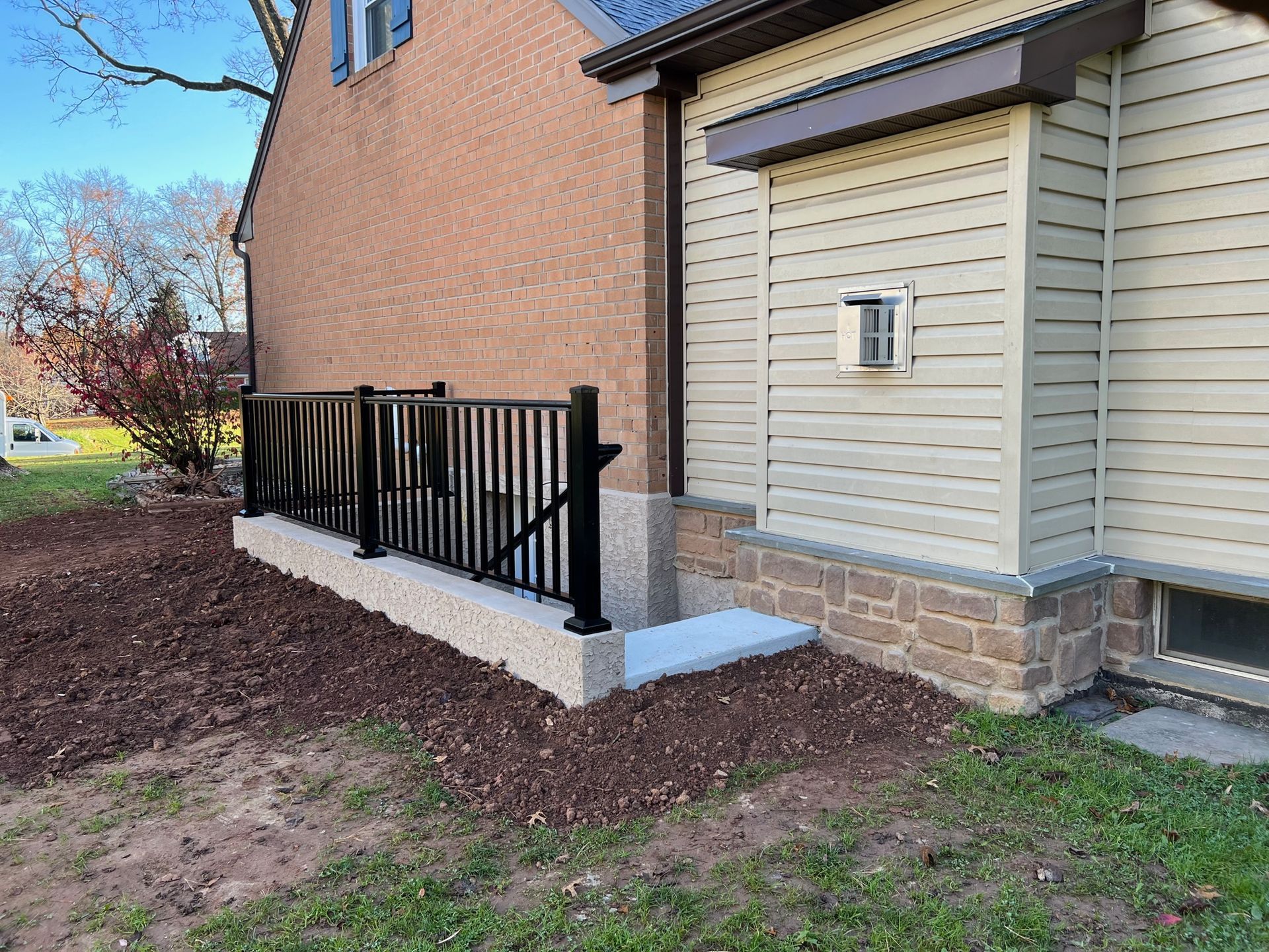 A black metal railing borders a concrete patio and walkway next to a house with brick and beige siding.