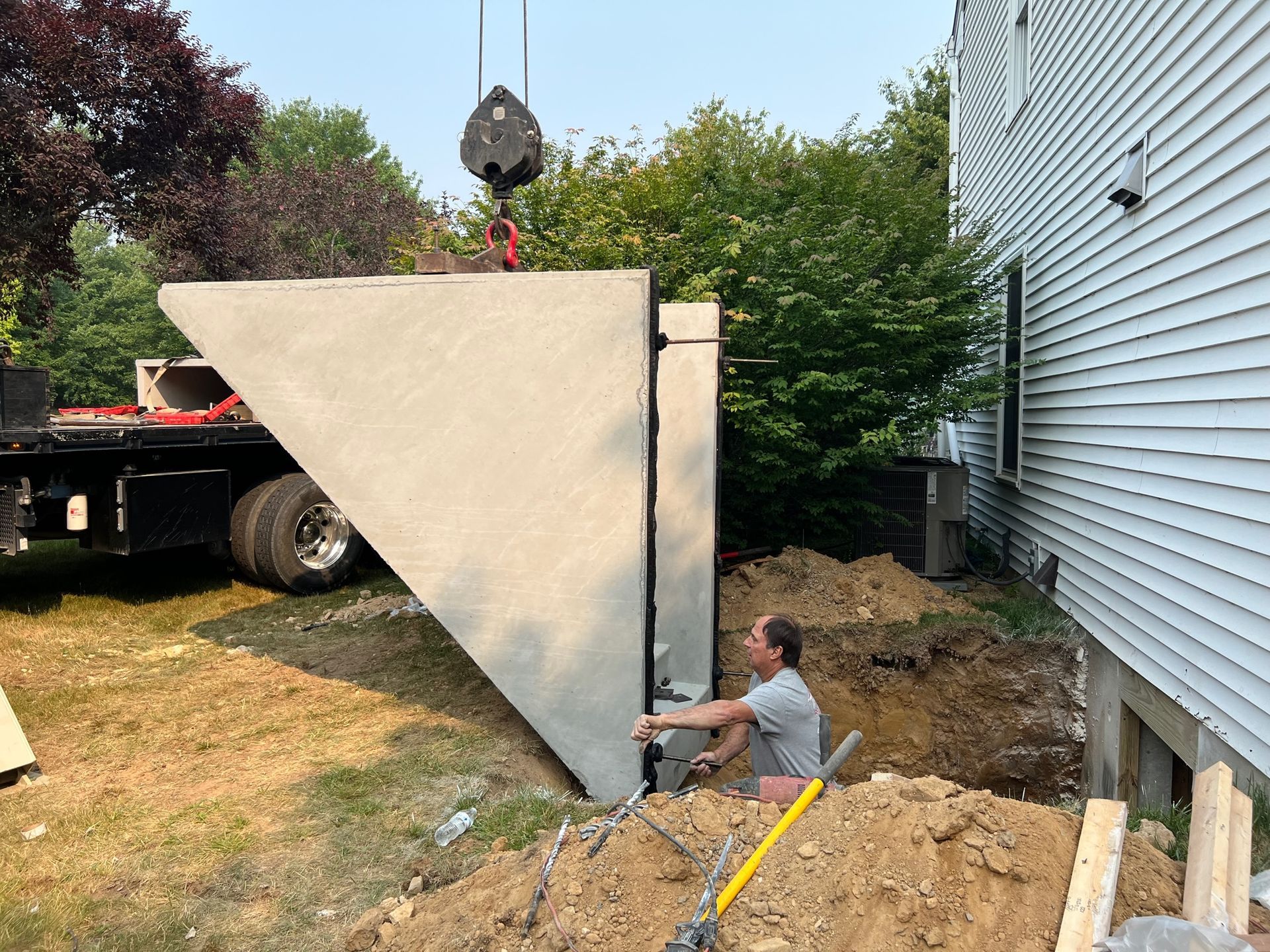 Worker installing a large concrete wall panel beside a house, lifted by a crane