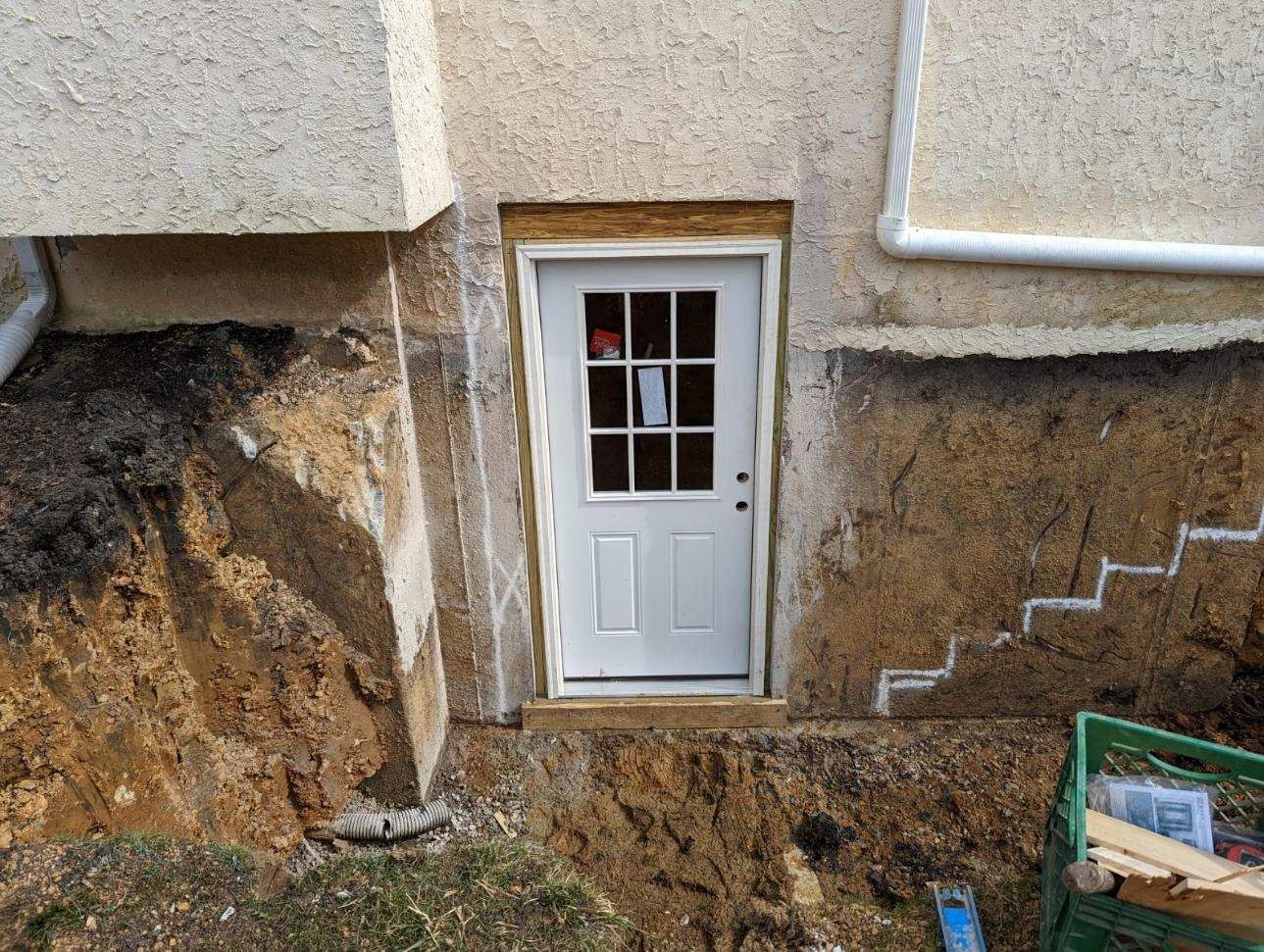 Basement door in a dirt excavation beside a house foundation, with stairs on the right.