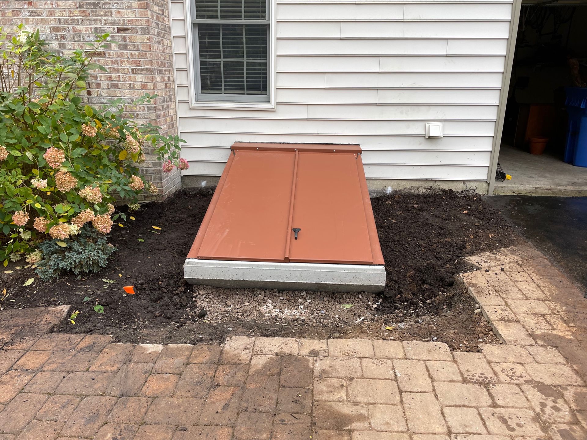 Exterior basement hatch with rust-colored cover beside a house and brick patio