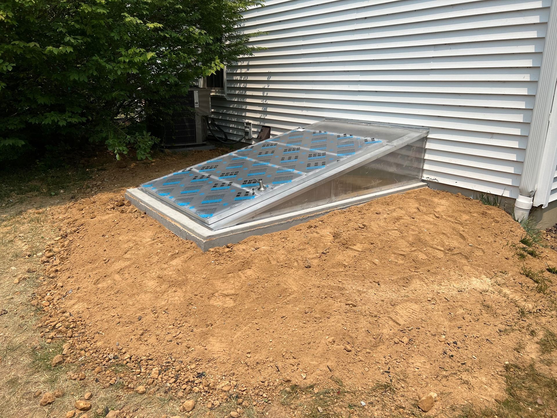 Sloped basement window well beside a house, bordered by dirt and trees