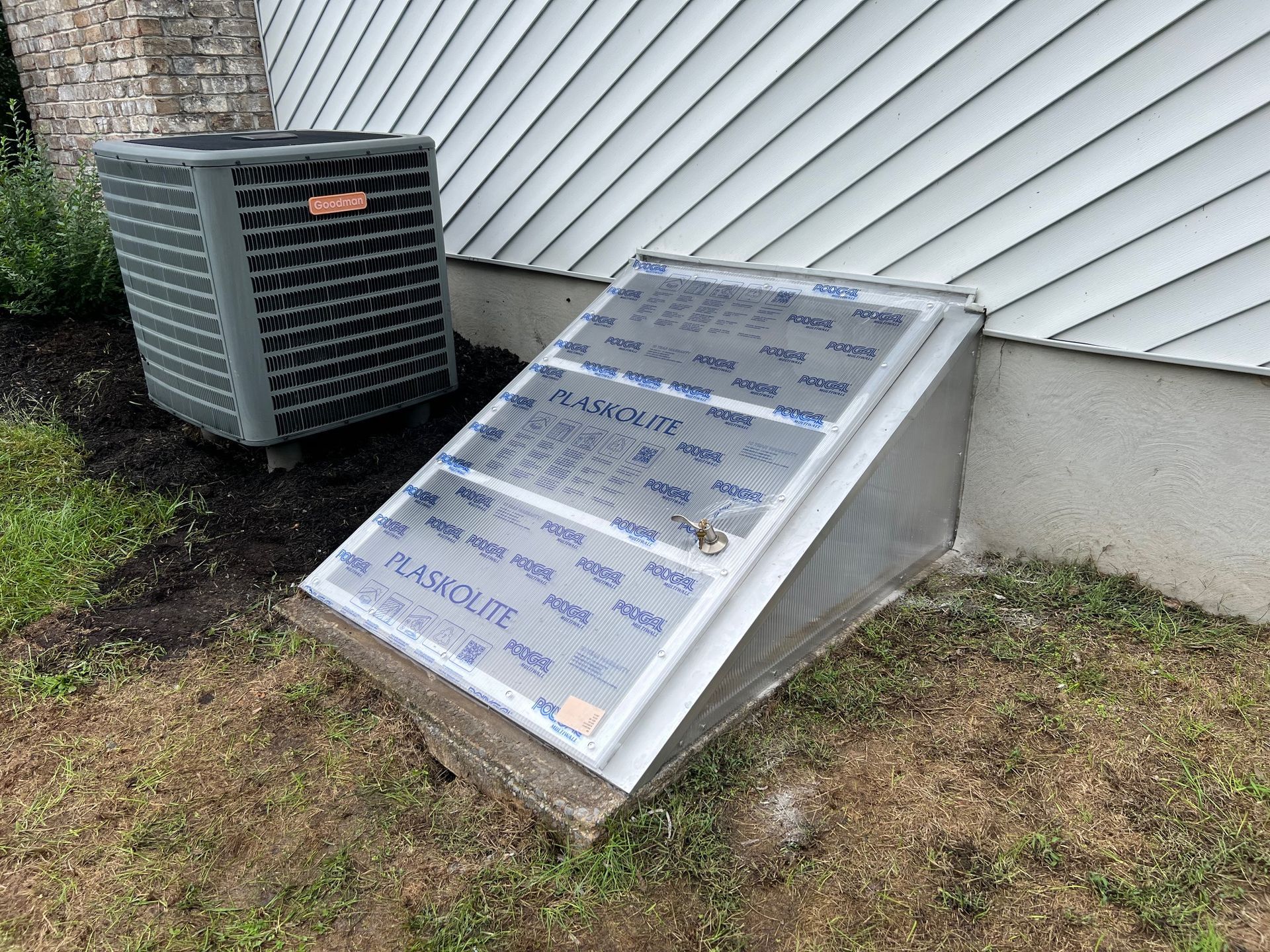 An air conditioner is sitting on the side of a house next to a window well.