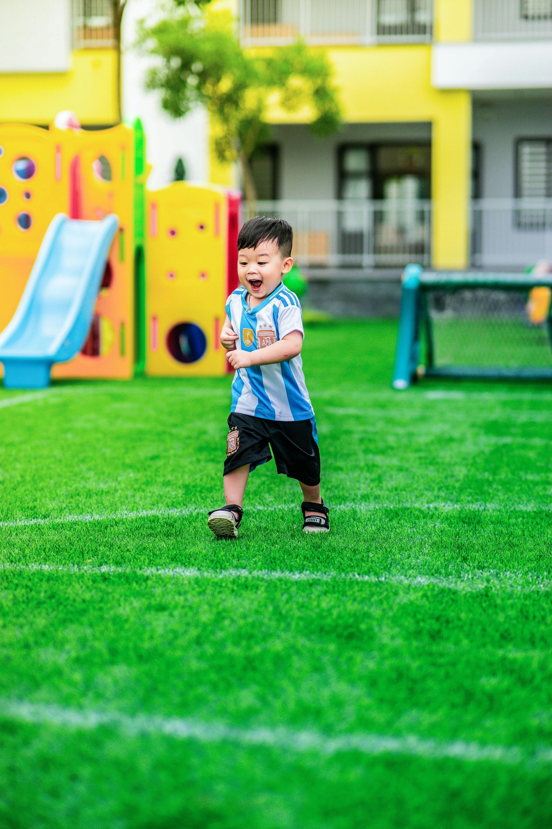 Child standing on a green playground lawn with slides and play equipment in the background